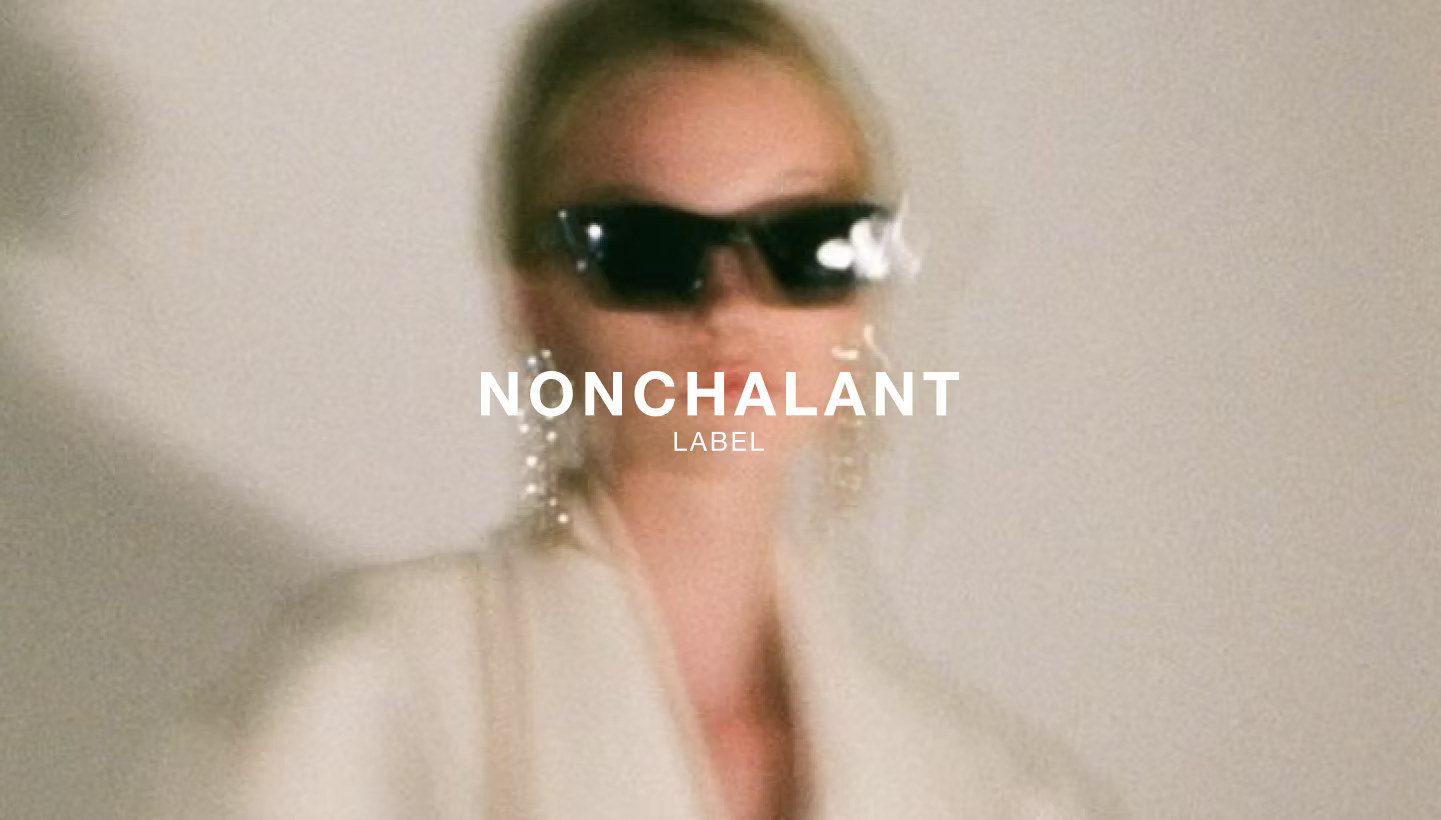 A woman in cream tailoring moving through frame, out of focus, under the Nonchalant Label mark