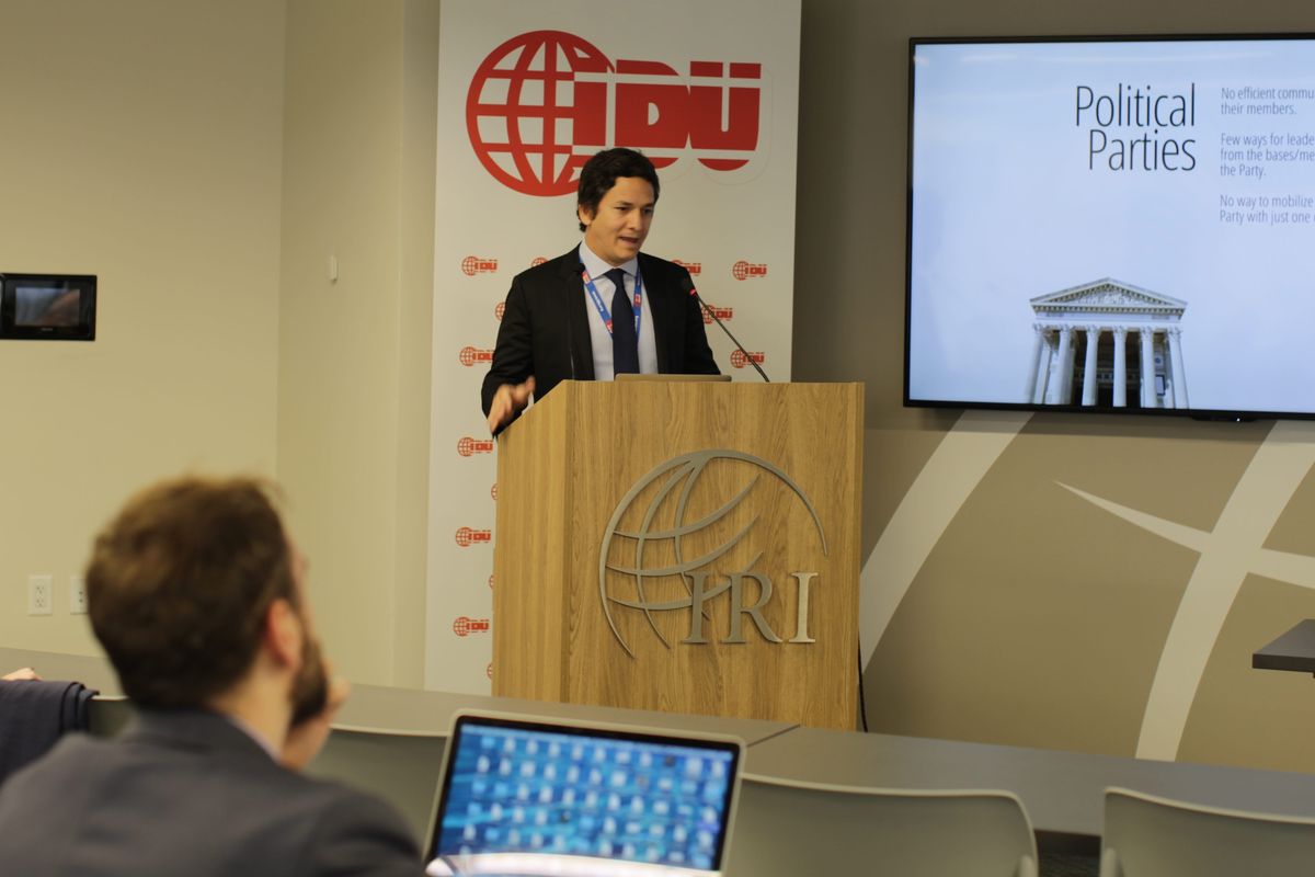 Ricardo De La Blanca Brigati at a lectern in front of an International Democrat Union backdrop, a slide on party communication beside him