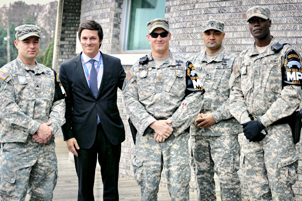 Ricardo De La Blanca Brigati in a suit, standing with United States Army personnel whose unit patches read Pan Mun Jom