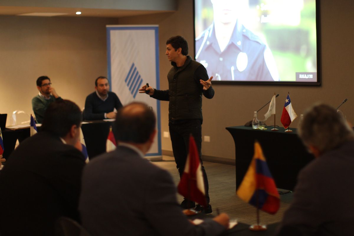 A man speaking to a seated panel, a Chilean flag on the table and a foundation banner behind